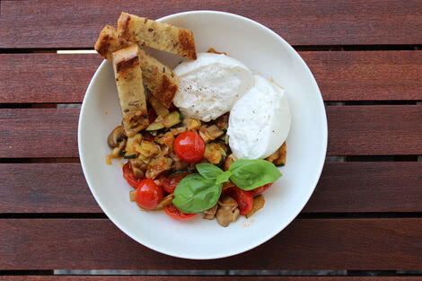 Auf dem Bild sind zwei pochierte Eier auf einer Gemüsemischung mit Tomaten und Pilzen, garniert mit frischem Basilikum und dazu geröstetes Brot zu sehen.
