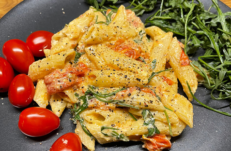 Auf dem Bild ist eine Portion Pasta mit Kirschtomaten und Rucola auf einem schwarzen Teller zu sehen.