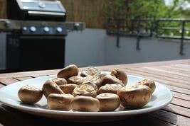 Auf dem Bild sind frische Champignons auf einem weißen Teller zu sehen, mit einem Grill im Hintergrund.