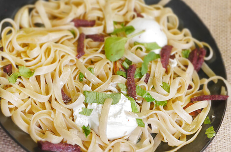 Auf dem Bild sind Bandnudeln mit Stückchen von Wurst und frischen Kräutern sowie einer Portion Sahne zu sehen.