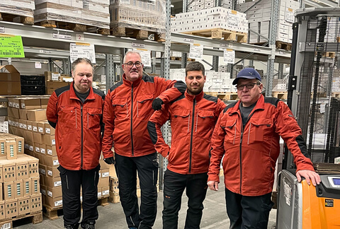 Auf dem Bild sind vier Männer in roten Arbeitsjacken in einem Lager zu sehen.