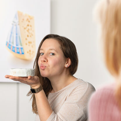 Auf dem Bild ist eine Frau zu sehen, die einen Käse in der Hand hält und sich mit einem spielerischen Gesichtsausdruck präsentiert.