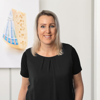 Auf dem Bild ist eine lächelnde Person in einem schwarzen Oberteil vor einem Hintergrund mit einem Käse zu sehen.