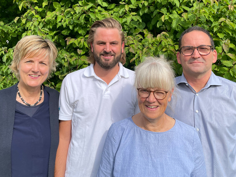 The image features four individuals standing before a lush green hedge, echoing the natural setting where our Allgäu region’s organic cheeses are crafted.