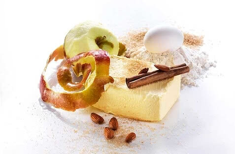 Auf dem Bild sind verschiedene Zutaten zu sehen, darunter Butter, Äpfel, Mehl, ein Ei, Zimtstangen und Mandeln.
