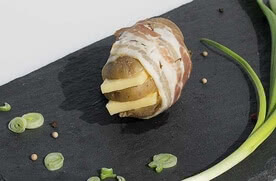 Auf dem Bild ist eine Kartoffel zu sehen, die mit Speck umwickelt ist und mit Käsestücken gefüllt ist, umgeben von einigen frischen Frühlingszwiebeln.