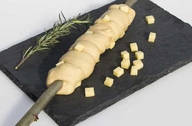 Auf dem Bild ist ein Stock zu sehen, der mit Teig umwickelt und mit Käsewürfeln versehen ist, dazu liegt ein Zweig Rosmarin und weitere Käsewürfel auf einem schwarzen Steinbrett.