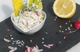 Das Bild zeigt eine Schüssel mit einem cremigen Salat oder Dip, dekoriert mit Salatblättern, Radieschen, Zitronenhälften und verschiedenen Gewürzen auf einer schwarzen Platte.