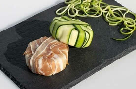 Auf dem Bild sind zwei kleine, kunstvoll angerichtete Speisen zu sehen: eine mit Schinken umwickelte Füllung und eine mit Zucchini umhüllte Füllung, die auf einem schwarzen Stein platziert sind, umgeben von spiralförmig geschnittenen Zucchini.