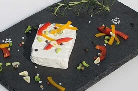 Auf dem Bild ist ein Stück Frischkäse dekoriert mit buntem Gemüse und Gewürzen auf einem schwarzen Servierbrett zu sehen.