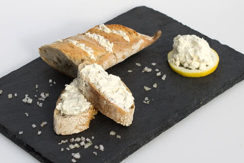 Auf dem Bild sind ein Stück Brot mit Aufstrich und eine Zitronenscheibe zu sehen, die auf einem schwarzen Steinbrett angerichtet sind.