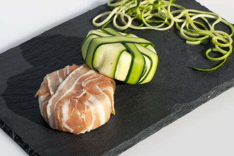 Auf dem Bild sind zwei kunstvoll dekorierte Häppchen zu sehen, eines aus Speck und das andere aus Zucchini, auf einem schwarzen Servierbrett.