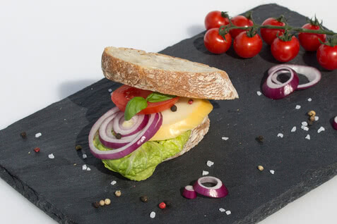 Auf dem Bild ist ein Sandwich mit frischem Gemüse, Käse und einer Brotscheibe zu sehen, das auf einem schwarzen Steinbrett präsentiert wird.