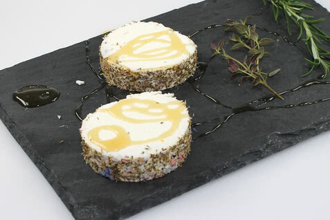 Auf dem Bild sind zwei runde Käseportionen mit Kräutern und Honig auf einem schwarzen Steinbrett zu sehen.